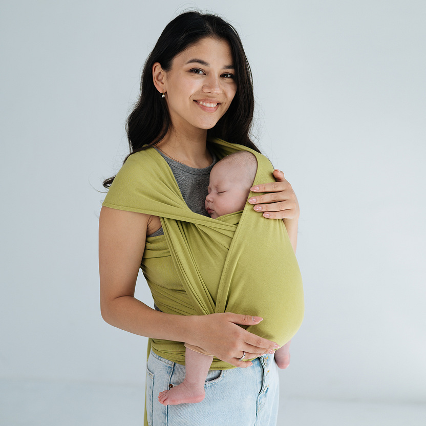 A mother wearing a light green modal stretchy wrap, holding her newborn baby.