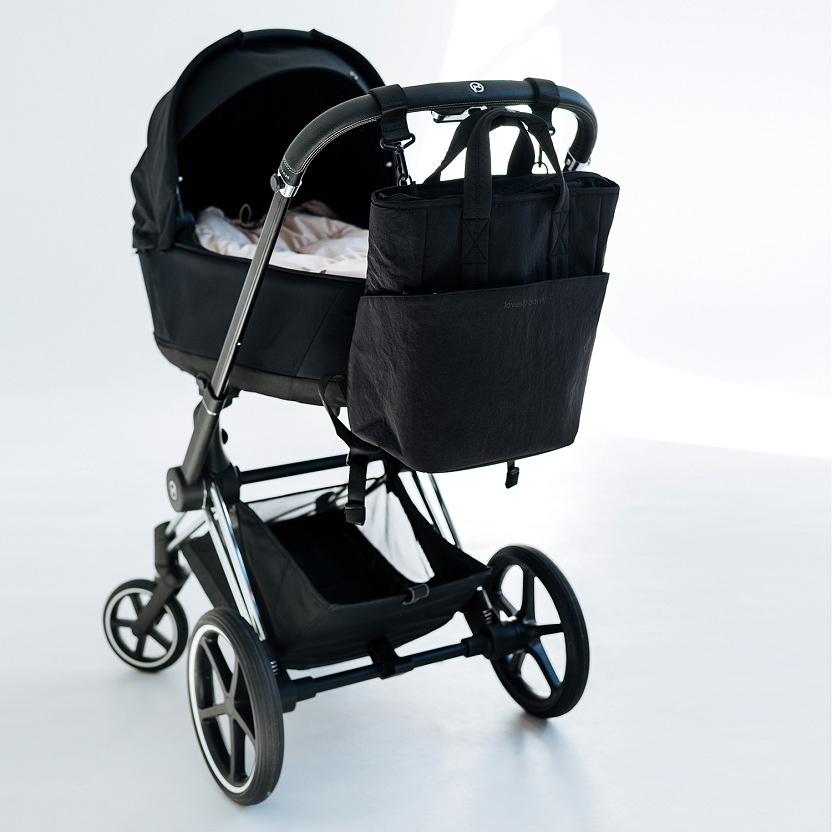 A black Love & Carry multifunctional diaper bag attached to the handlebar of a baby stroller. Convenient stroller accessory for organized storage on the go.